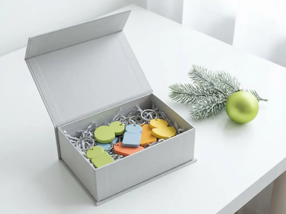 Scatola regalo aperta con portachiavi personalizzati per Natale in PVC colorato, ramo di abete e pallina verde