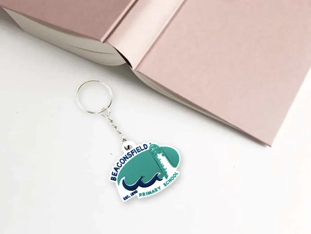 Portachiavi in PVC 2D a scudo con il logo di una scuola, appoggiato a un libro rosa